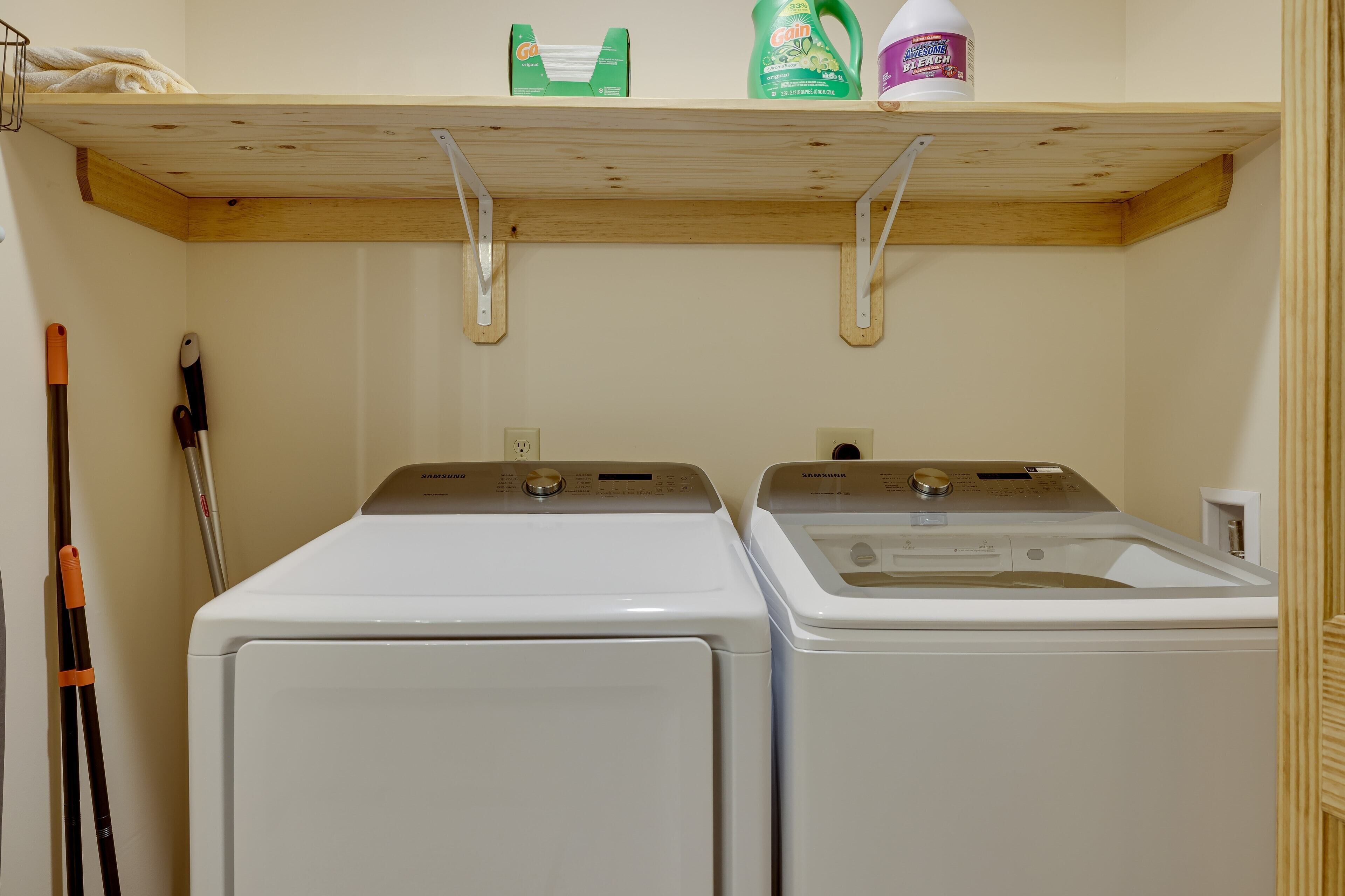 Two white front-loading washers with a wooden shelf above them holding cleaning supplies.