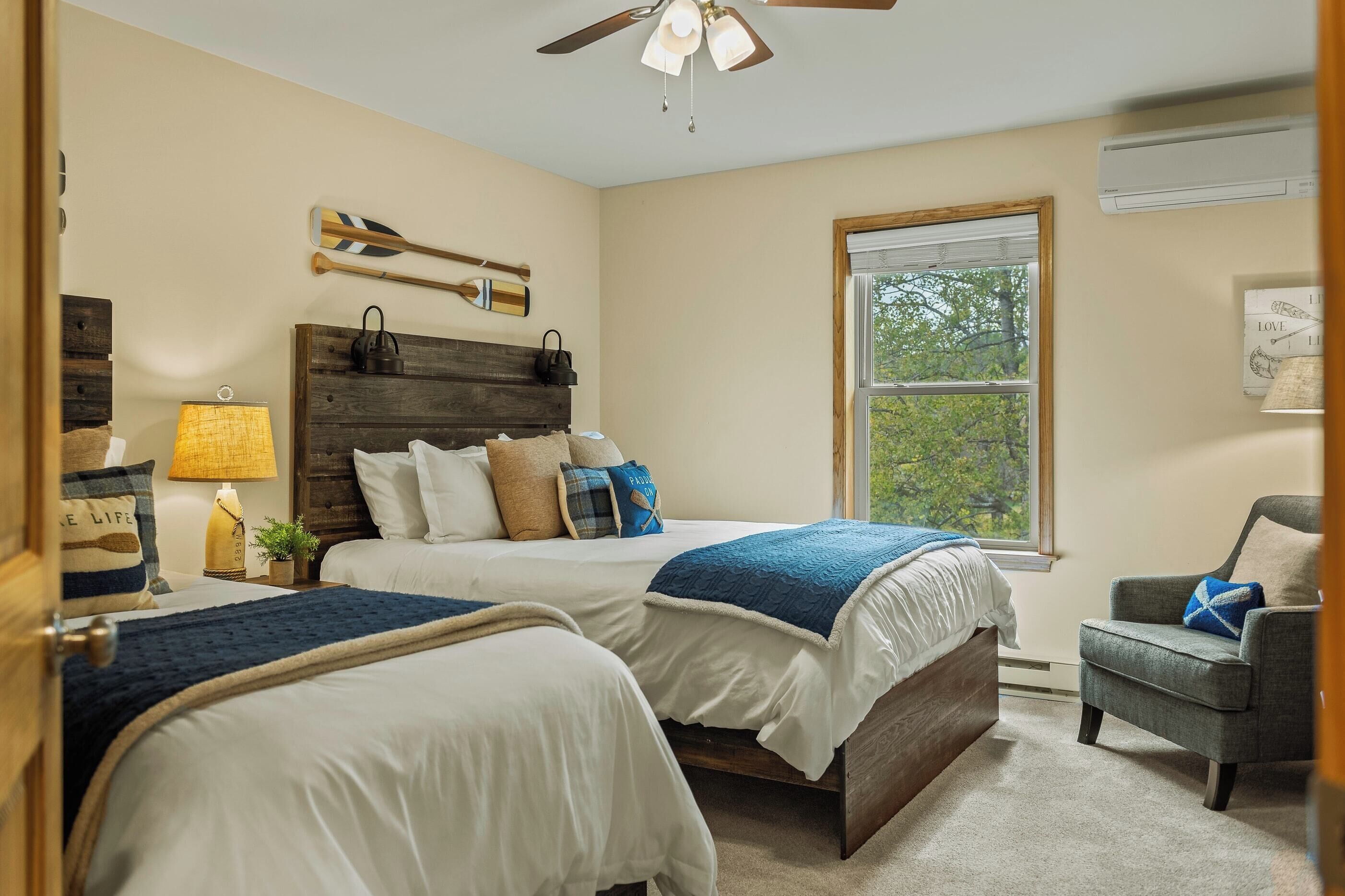 Une chambre avec deux lits, une tête de lit en bois, une fenêtre avec vue, un ventilateur de plafond et une chaise.