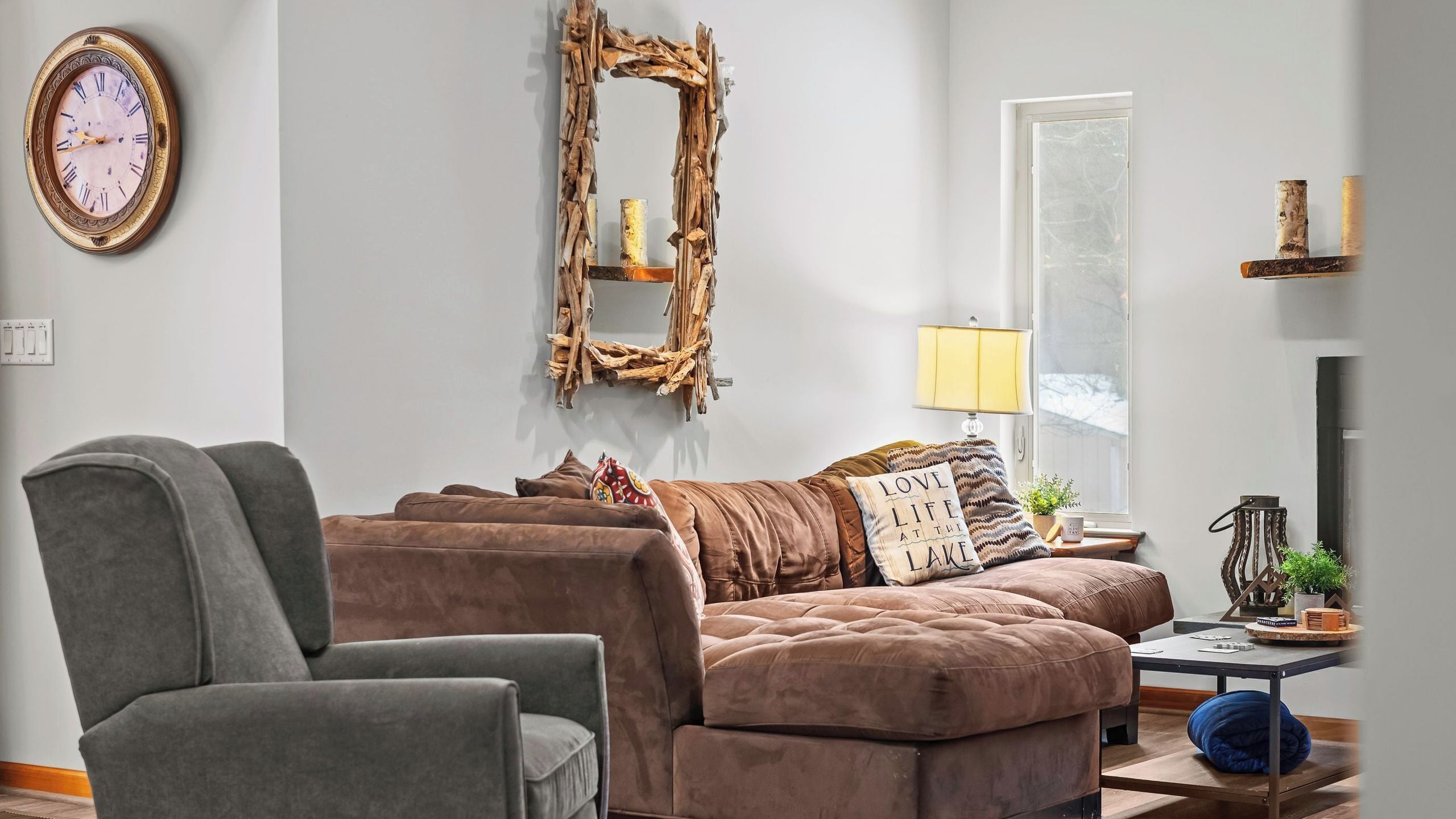 A living room with a brown sofa, a grey armchair, a wooden wall decor, a lamp, and a clock.