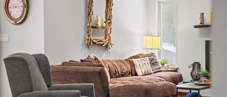 A living room with a brown sofa, a grey armchair, a wooden wall decor, a lamp, and a clock.