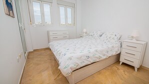 Um quarto com cama, duas mesas de cabeceira, um criado-mudo, uma janela e uma porta.