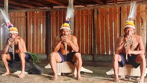 Three individuals seated on wooden benches in a rustic setting with wooden walls and a thatched roof.