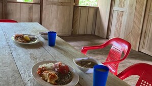 A wooden table with plates of food and blue cups, surrounded by red plastic chairs in a rustic setting with wooden walls and a thatched roof.