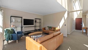 A spacious living room with a large sectional sofa, a blue armchair, and a coffee table. There is a black shelving unit with books and decorative items, and a flat-screen TV mounted on the wall. - Fresh on Fletcher - Taupo Holiday Home (Taupo)