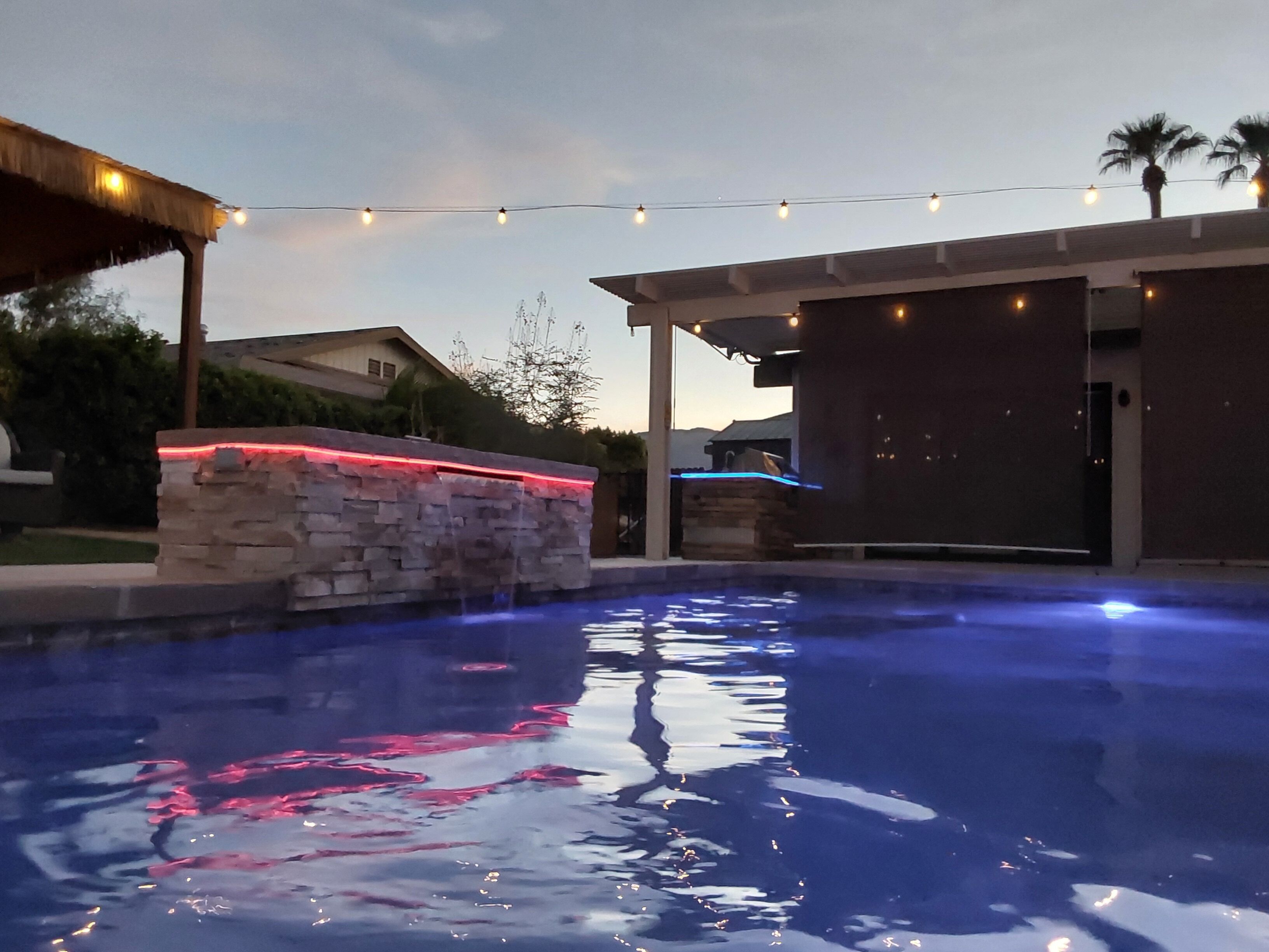 An outdoor swimming pool with a stone wall and a covered area with string lights.