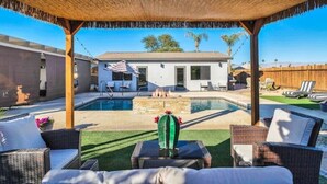 A covered outdoor area with a pool, lounge chairs, and a house in the background.