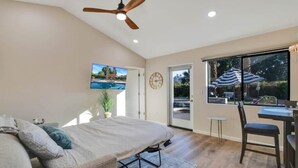 A bedroom with a bed, a ceiling fan, a TV, a window with a view of the outdoors, and a dining area with a table and chairs.