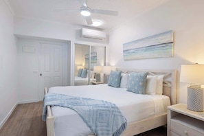A bedroom with a bed, bedside table, lamp, air conditioning unit, ceiling fan, and a painting on the wall.