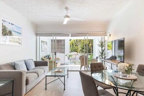 A modern living room with a sofa, coffee table, and dining table, large windows, and a ceiling fan.