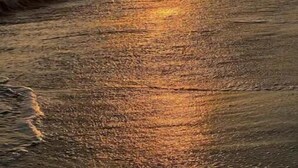 Auringonlasku rannan yllä, taustalla näkyvät aallot ja kivikkoiset kalliot.