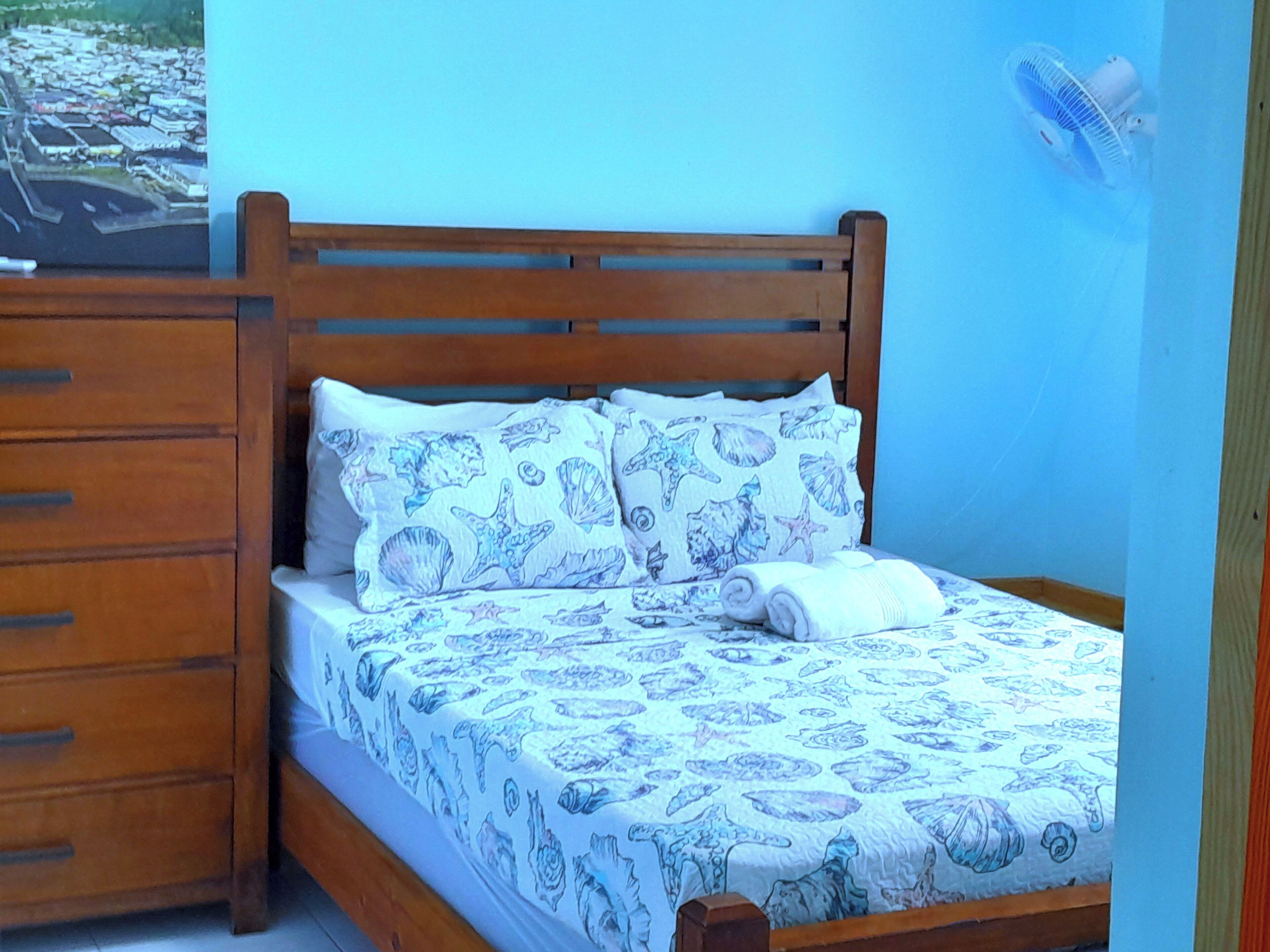 A bed with a blue and white patterned bedspread, wooden headboard, and two pillows with a sea-themed design.