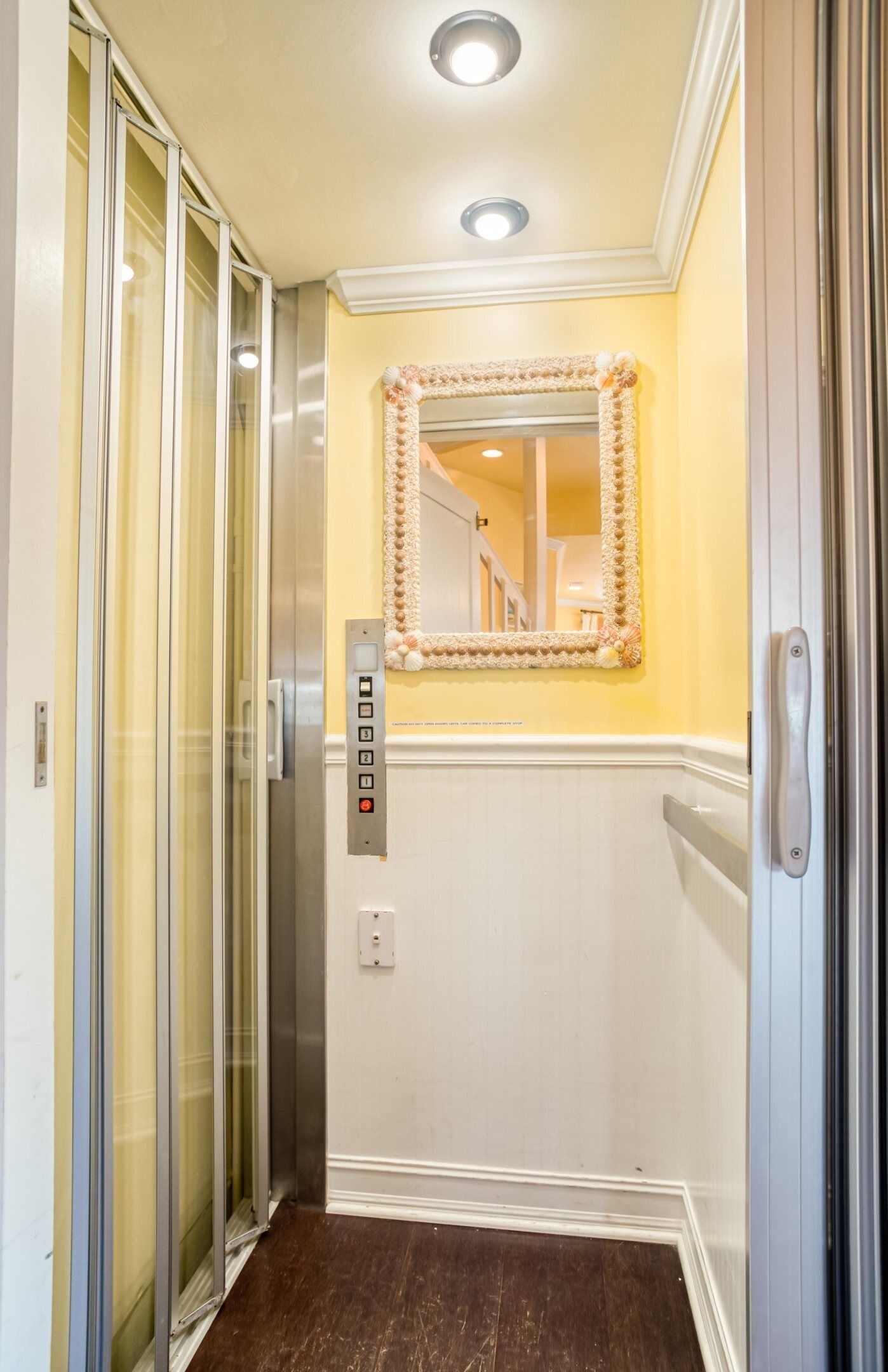 A narrow elevator with a mirror and control panel, white walls, and a wooden floor.