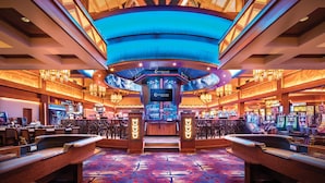 Interior de un casino con máquinas tragamonedas, una zona de bar y una gran pantalla digital.