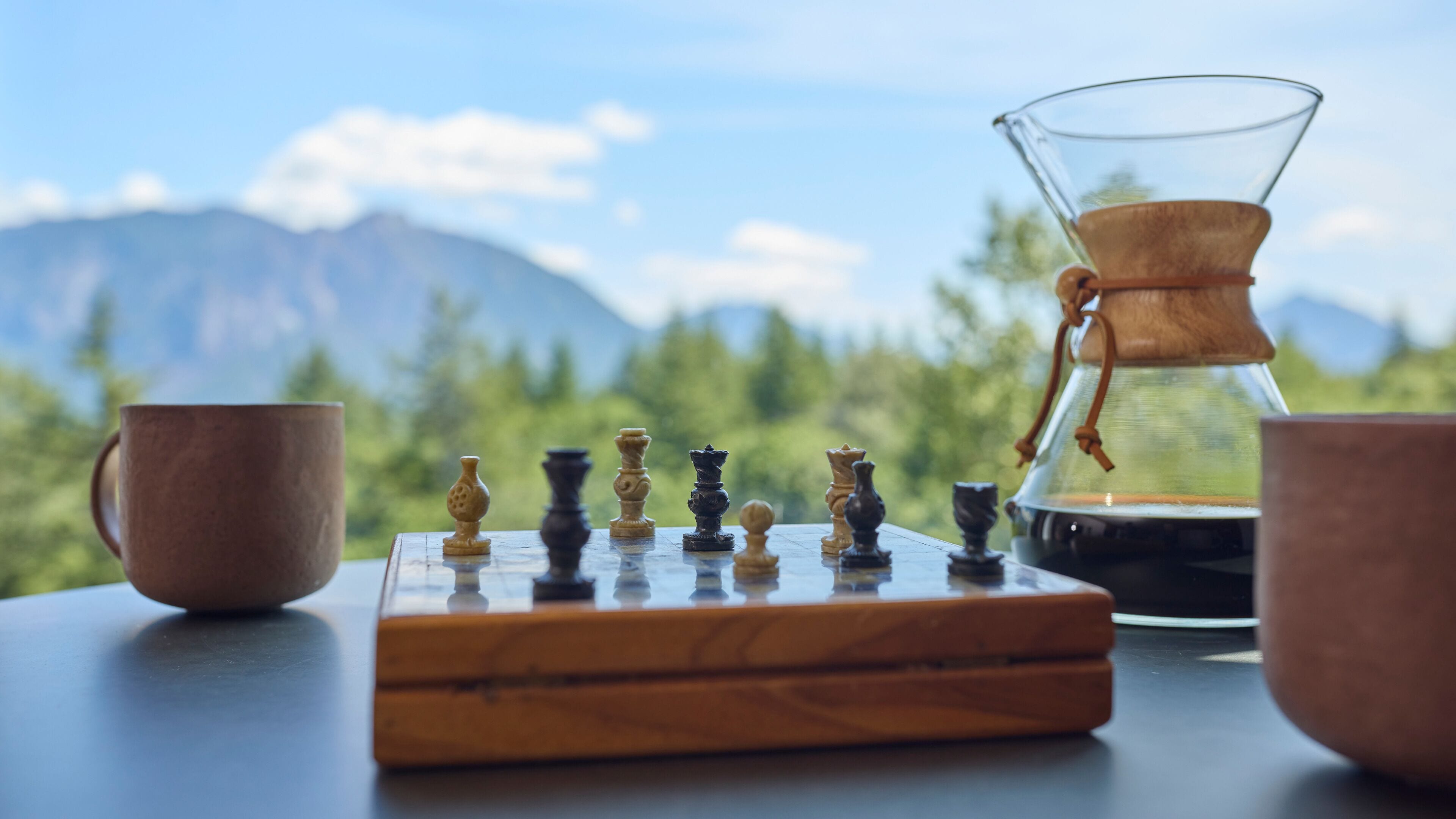A chessboard with pieces, a coffee pot, and two cups on a table with a mountain view.