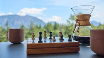A chessboard with pieces, a coffee pot, and two cups on a table with a mountain view.