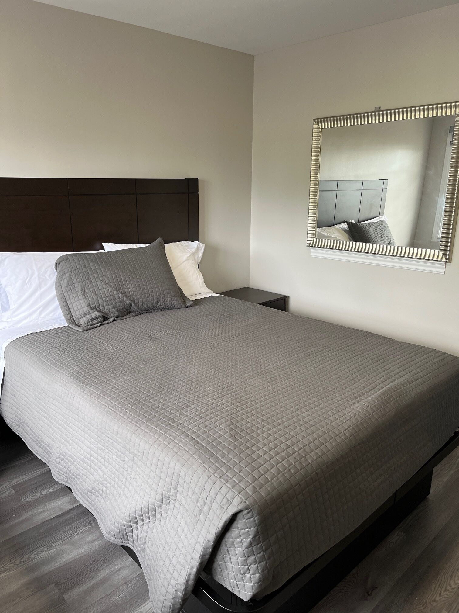 A neatly made bed with a quilted grey bedspread, a dark headboard, and a bedside table with a lamp.