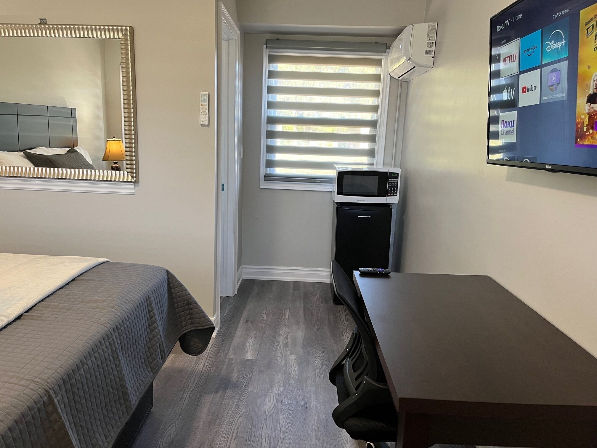 A compact hotel room with a bed, a desk, a chair, a microwave, and a TV displaying streaming services.