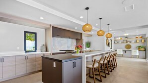 A modern kitchen with a central island, bar stools, pendant lighting, and a view of greenery outside. - Goldenview | 4BR Ocean, City & Bay Escape + Spa (San Diego)