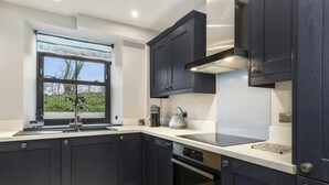 A modern kitchen with dark cabinetry, a stainless steel range hood, a built-in oven, and a window with a view of greenery. - Quarry House, Aberporth (Aberporth)