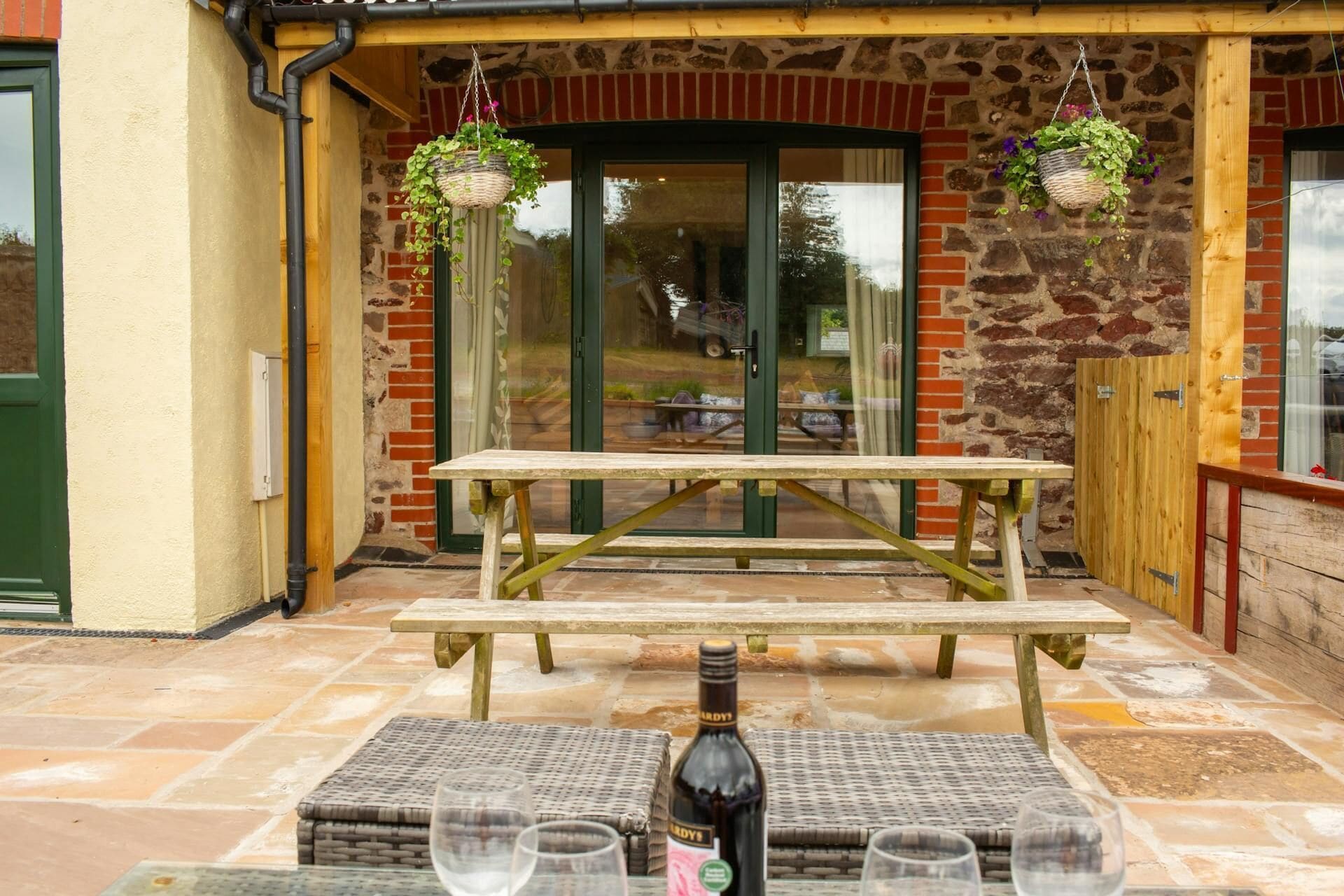 Un patio al aire libre con una mesa y bancos de madera, una botella de vino y dos macetas colgantes con plantas.