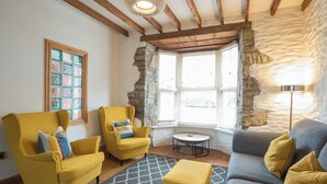 Un salon avec des fauteuils jaunes, un canapé gris, un tapis à motifs, une table ronde et un mur en pierre.