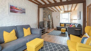 Un salon comprenant un canapé, un pouf et des fauteuils, une cheminée et un mur en pierre.