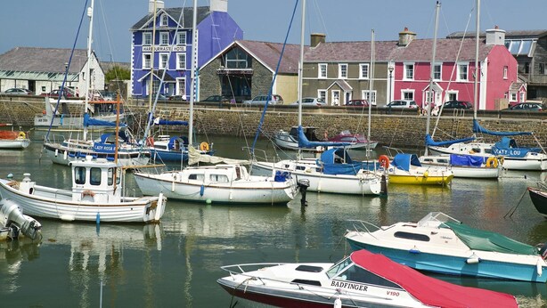 A harbor with various boats docked, a blue building labeled 'Harbourmaster Hotel', and a stone wall in the background.