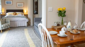 A hotel room with a bed, bedside table, chair, and a dining area with a table, chairs, and a vase of flowers.