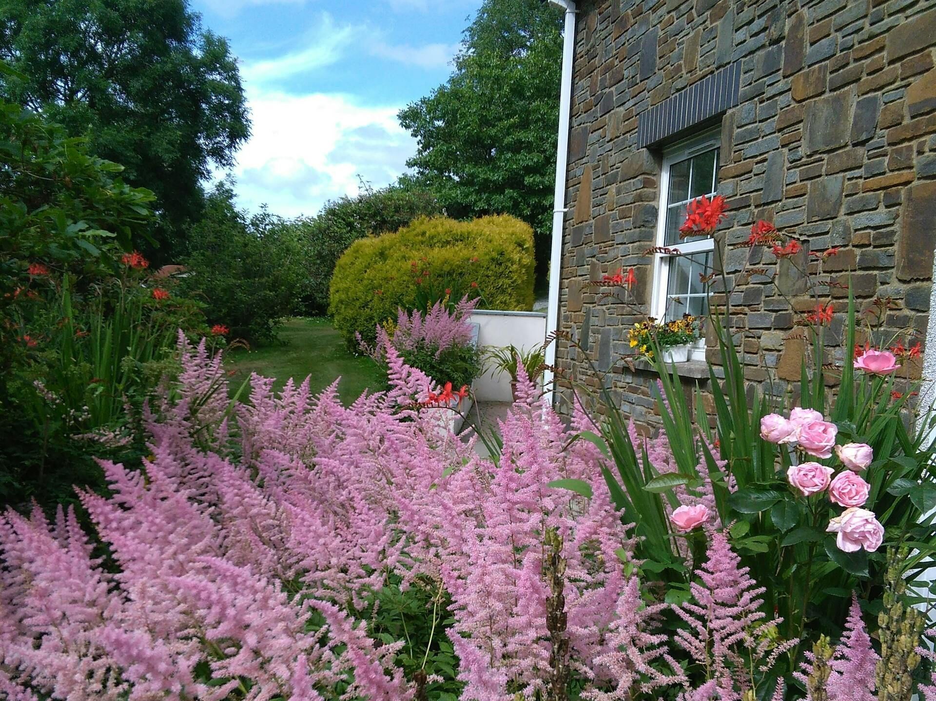 A stone building with a window displaying red flowers, surrounded by lush greenery and pink flowering plants.