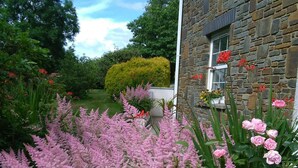 Un bâtiment en pierre avec une fenêtre ornée de fleurs rouges, entouré d’une végétation luxuriante et de plantes à fleurs roses.
