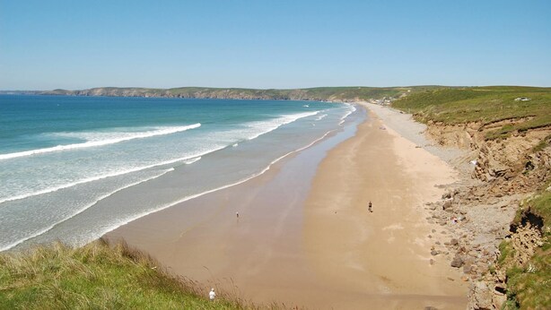 A coastal beach with clear skies, a sandy shore, and grassy hills in the background.