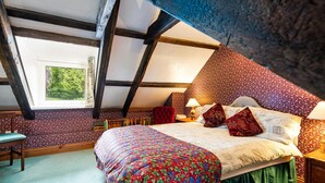 Una camera da letto con soffitto spiovente, un letto con lenzuola a motivi floreali, una poltrona rossa e un comodino con una lampada.