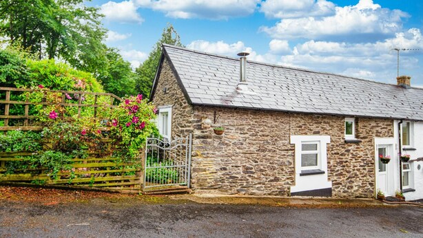 Un cottage in pietra con tetto spiovente, una recinzione in legno e un giardino fiorito di fiori rosa.