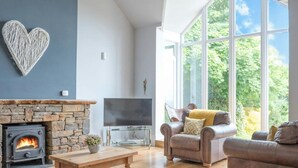 A modern living room with a stone fireplace, a flat-screen TV, leather sofas, and large windows offering a view of greenery. - Stella Maris, Freshwater East (Freshwater East)