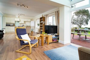A modern living room with a TV, wooden furniture, a kitchen in the background, and a view of the outdoors. - Safe Haven, Aberporth (Aberporth)