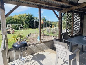 An outdoor patio area with a grill, chairs, and a view of a garden with a swing set and climbing frame.