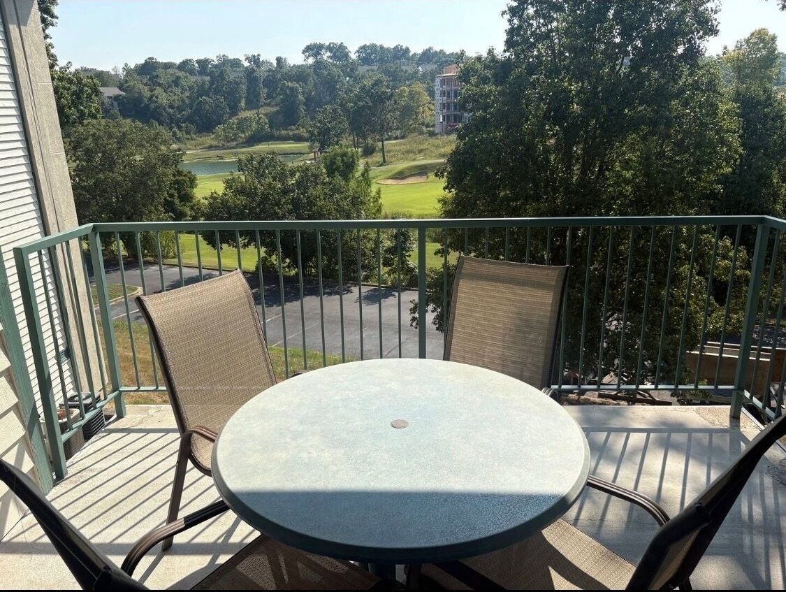 A balcony with a round table and two chairs, overlooking a golf course and trees.