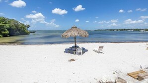 Een strand met een rieten parasol, witte ligstoelen en een helder uitzicht op de oceaan.