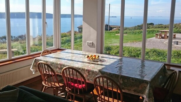 Un espace repas comprenant une table et des chaises, de grandes fenêtres offrant une vue sur un lac et la verdure environnante.