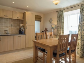 A kitchen with wooden cabinets, a dining table with chairs, a clock on the wall, and a window with curtains. - St Brides View, Solva (Solva)
