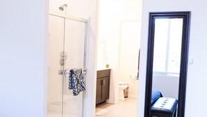 Une chambre comprenant un lit, un banc, une cabine de douche en verre, une armoire et un petit balcon avec une chaise.
