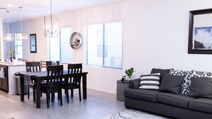 Un salon moderne avec un canapé gris, une table à manger avec des chaises et une grande fenêtre équipée de stores.