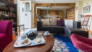 A cozy living room with a sofa, a coffee table with tea and cake, a bookshelf, and a kitchen visible through an open door. - The Stables, Newgale (Llandeloy)