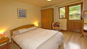 Een slaapkamer met een bed, een houten garderobe, een stoel en een raam met uitzicht op groen.