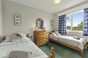 A bedroom with two single beds, a wooden dresser, a mirror, and a window with curtains. - Puffins Nest, Tenby - Two Bedroom Cottage, Sleeps 4 (Tenby)