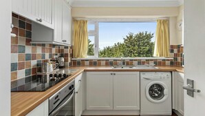 A compact kitchen with a built-in oven, a washing machine, and a window with curtains. - Puffins Nest, Tenby - Two Bedroom Cottage, Sleeps 4 (Tenby)