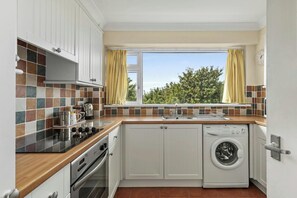 A compact kitchen with a built-in oven, a washing machine, and a window with curtains. - Puffins Nest, Tenby - Two Bedroom Cottage, Sleeps 4 (Tenby)