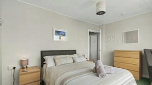 A bedroom with a bed, bedside tables, a lamp, a hanging light fixture, a door, and a chair. - Puffins Nest, Tenby - Two Bedroom Cottage, Sleeps 4 (Tenby)