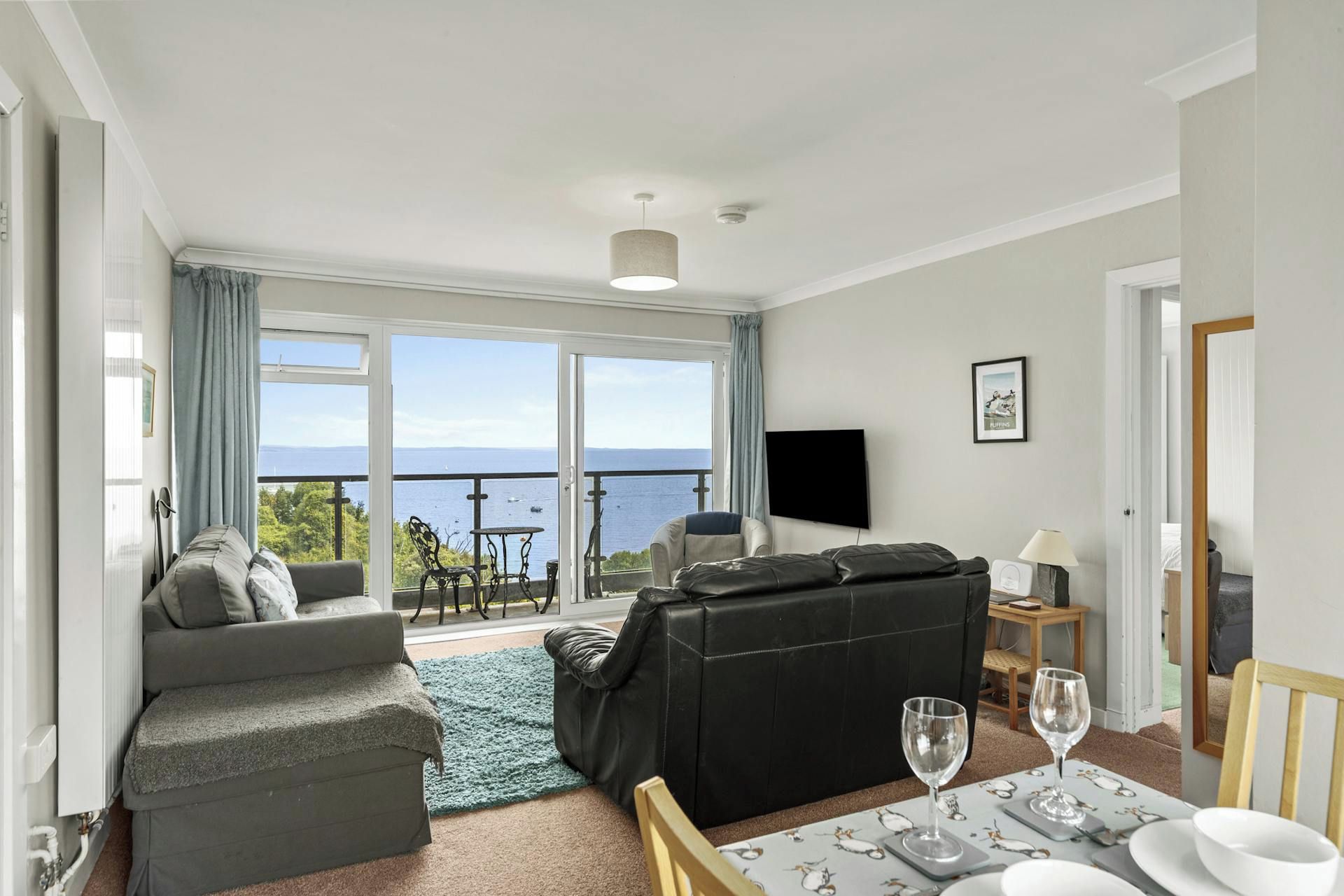 A living room with a sofa, a black leather couch, a dining table set with wine glasses, and a view of the sea through large windows.
