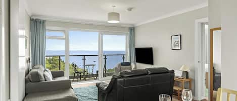En stue med sofa, lædersofa i sort, spisebord dækket med vinglas og havudsigt gennem store vinduer.